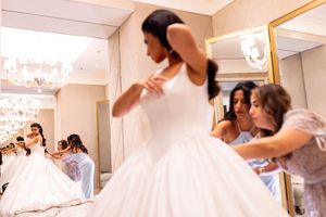 Bridesmaids helping bride adjust her white ball gown wedding dress in a bridal boutique fitting room.