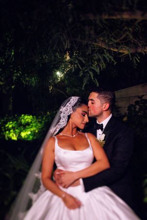 Groom in black tuxedo kisses bride wearing lace veil in a lush garden at night.