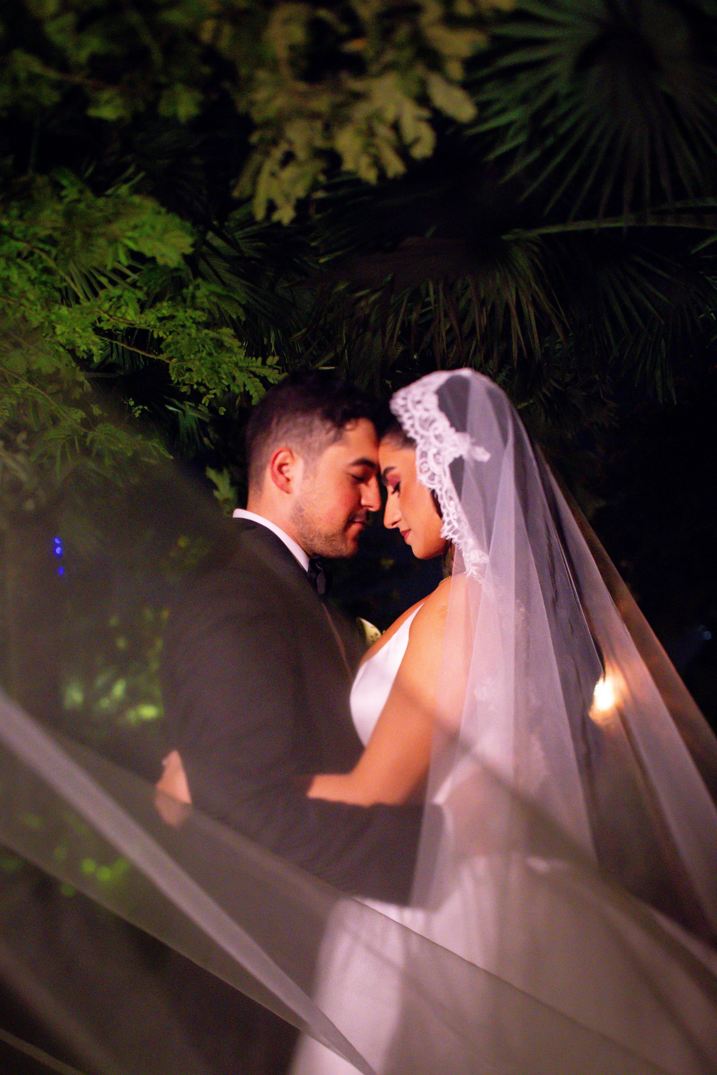 Bride and groom share a romantic moment under lush green foliage at night, her lace veil flowing beautifully.