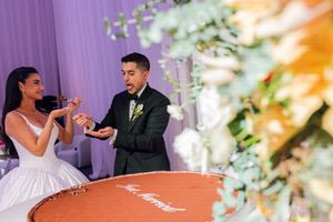Bride and groom cutting their wedding cake at elegant reception with purple backdrop and floral decor.