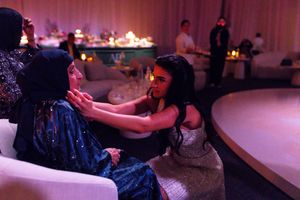 Two women in elegant gowns share an intimate moment at a glamorous evening event with soft pink ambient lighting.
