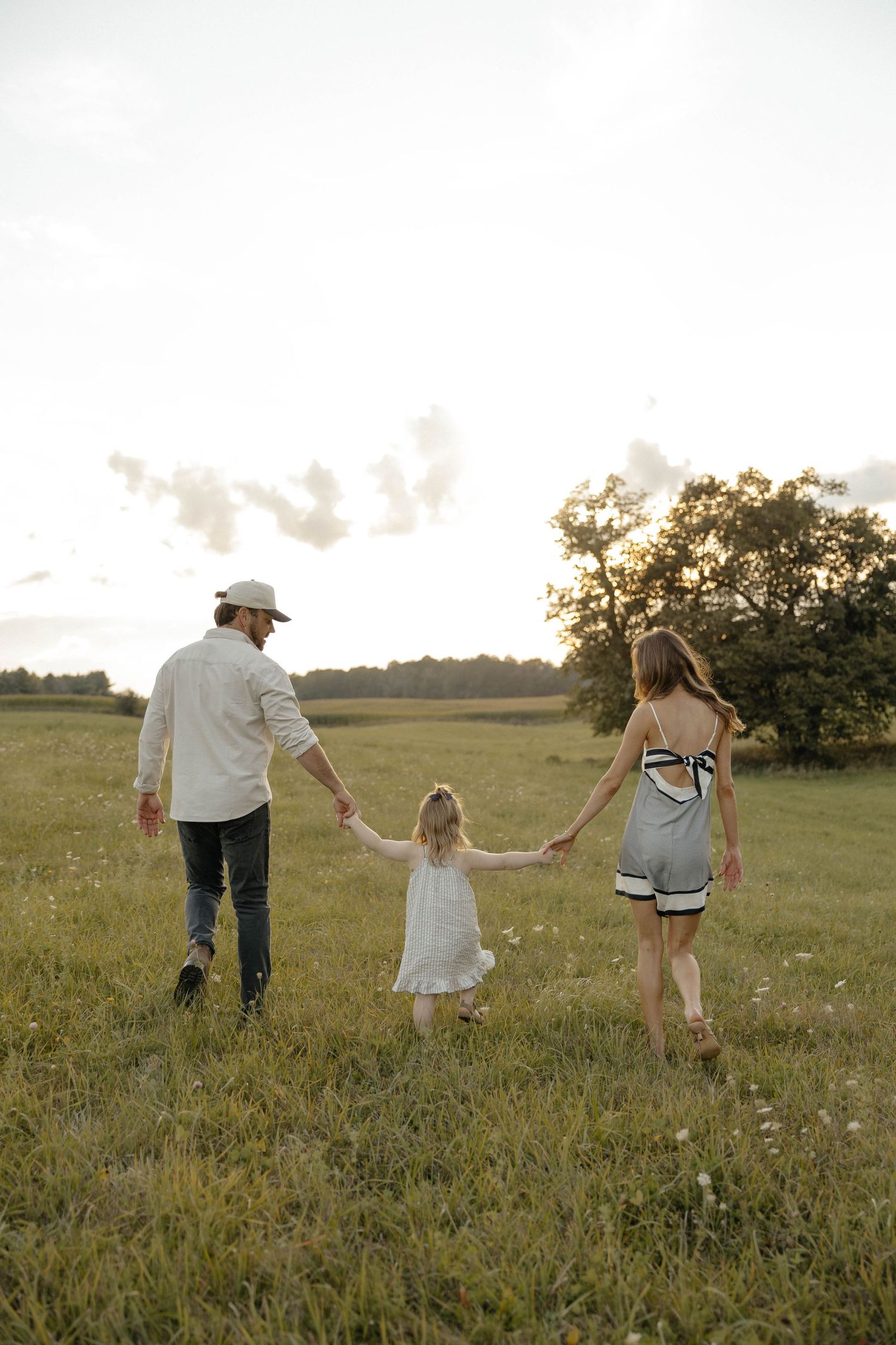 Family of three holding hands walking through a green meadow at golden hour, viewed from behind.