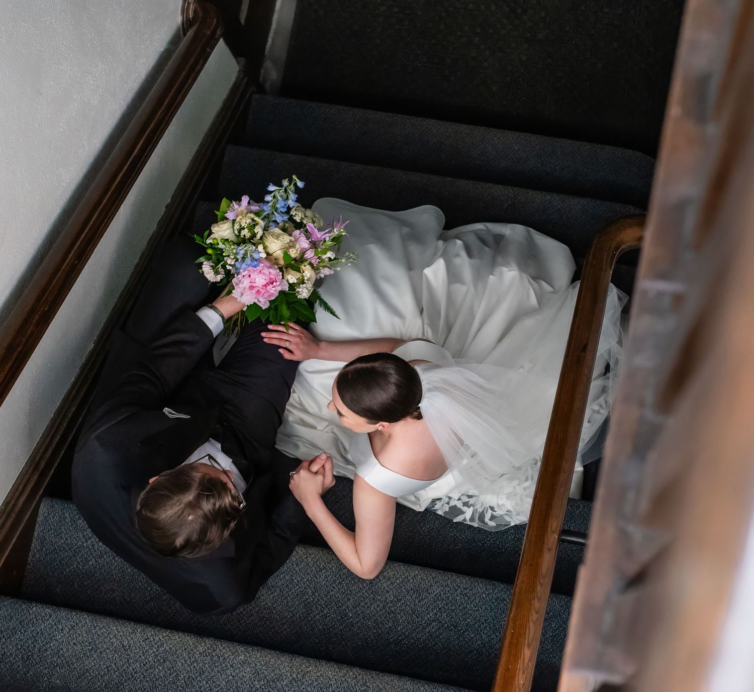 Aerial view of bride and groom sitting on carpeted stairs, bride holding colorful bouquet on their wedding day.