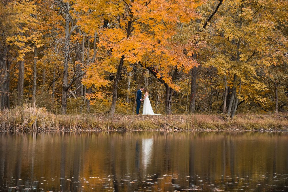 Couple embracing by a reflective pond surrounded by vibrant autumn foliage in orange and yellow hues.