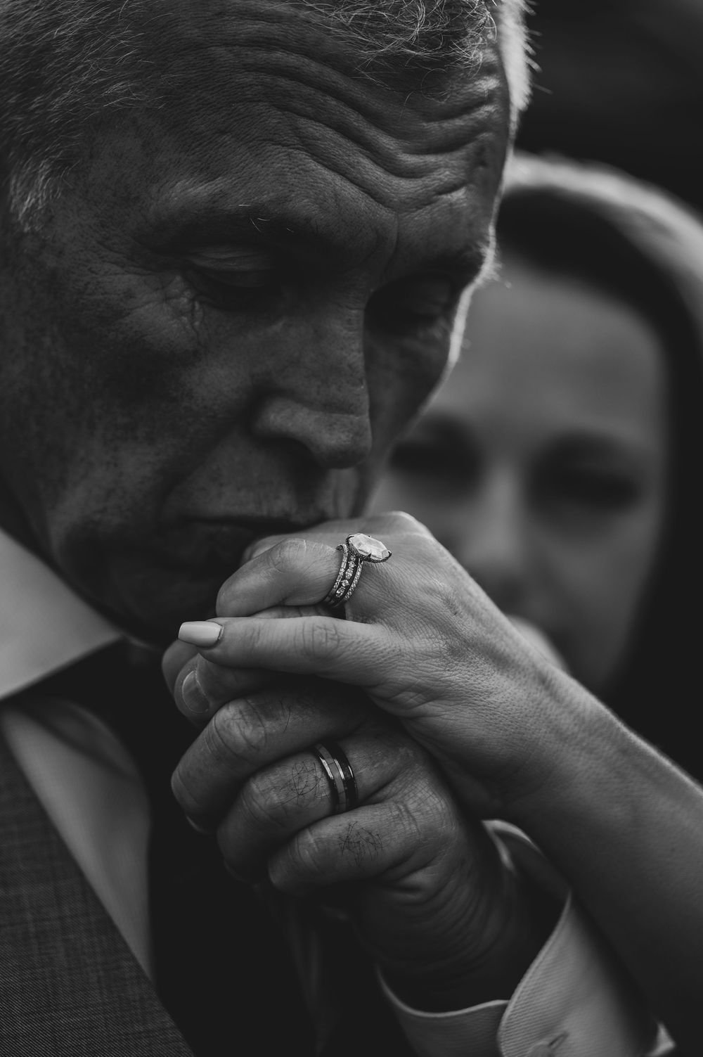 Black and white photo of an older man kissing a woman's hand with wedding rings visible at a formal event.