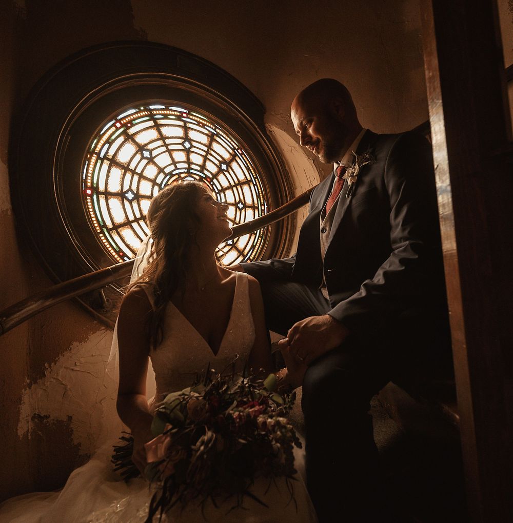 Bride and groom share a romantic moment on a staircase, backlit by a stunning circular window.