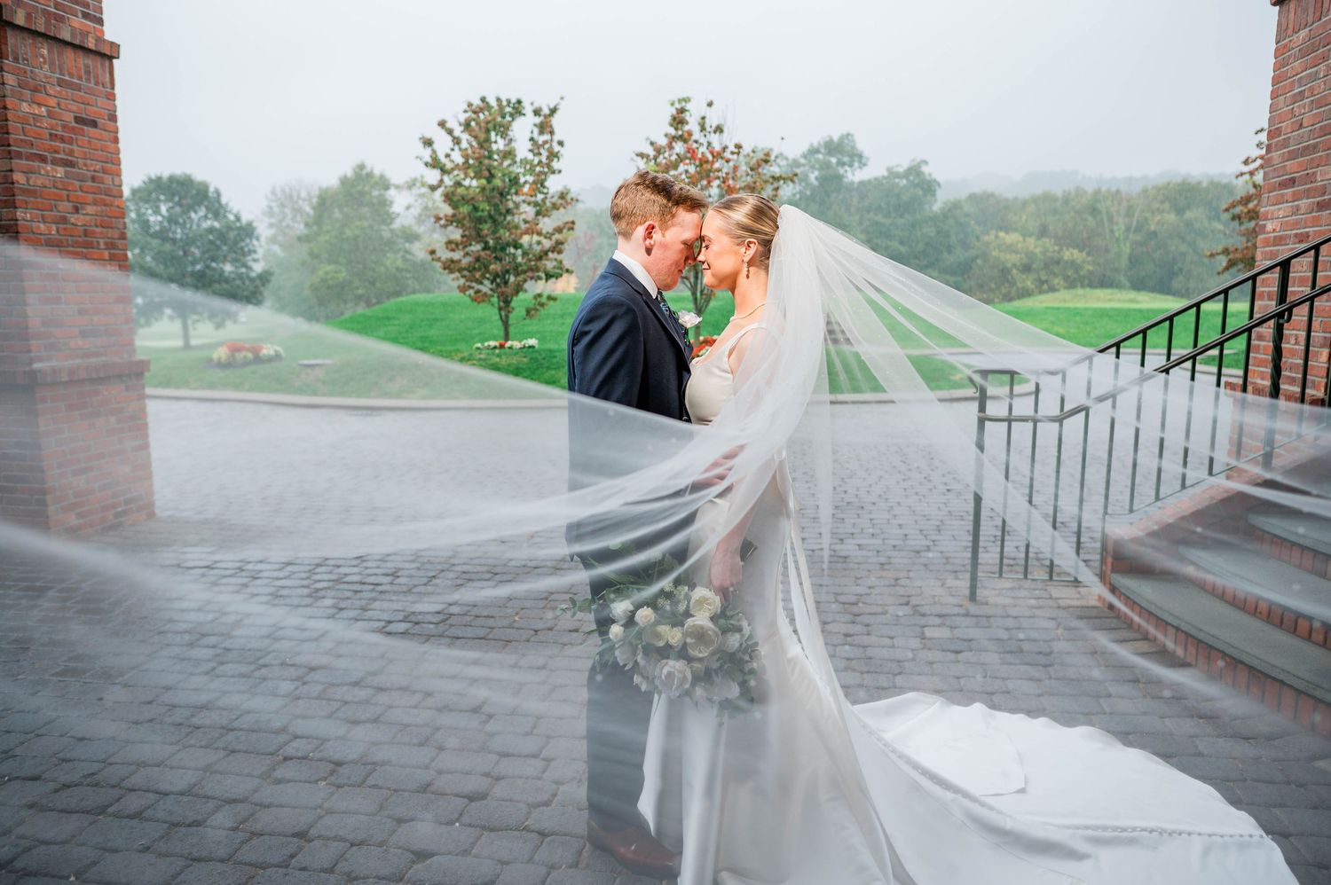 Bride and groom share romantic moment outdoors, wedding veil flowing dramatically in the wind on a misty day at Great River Golf Course