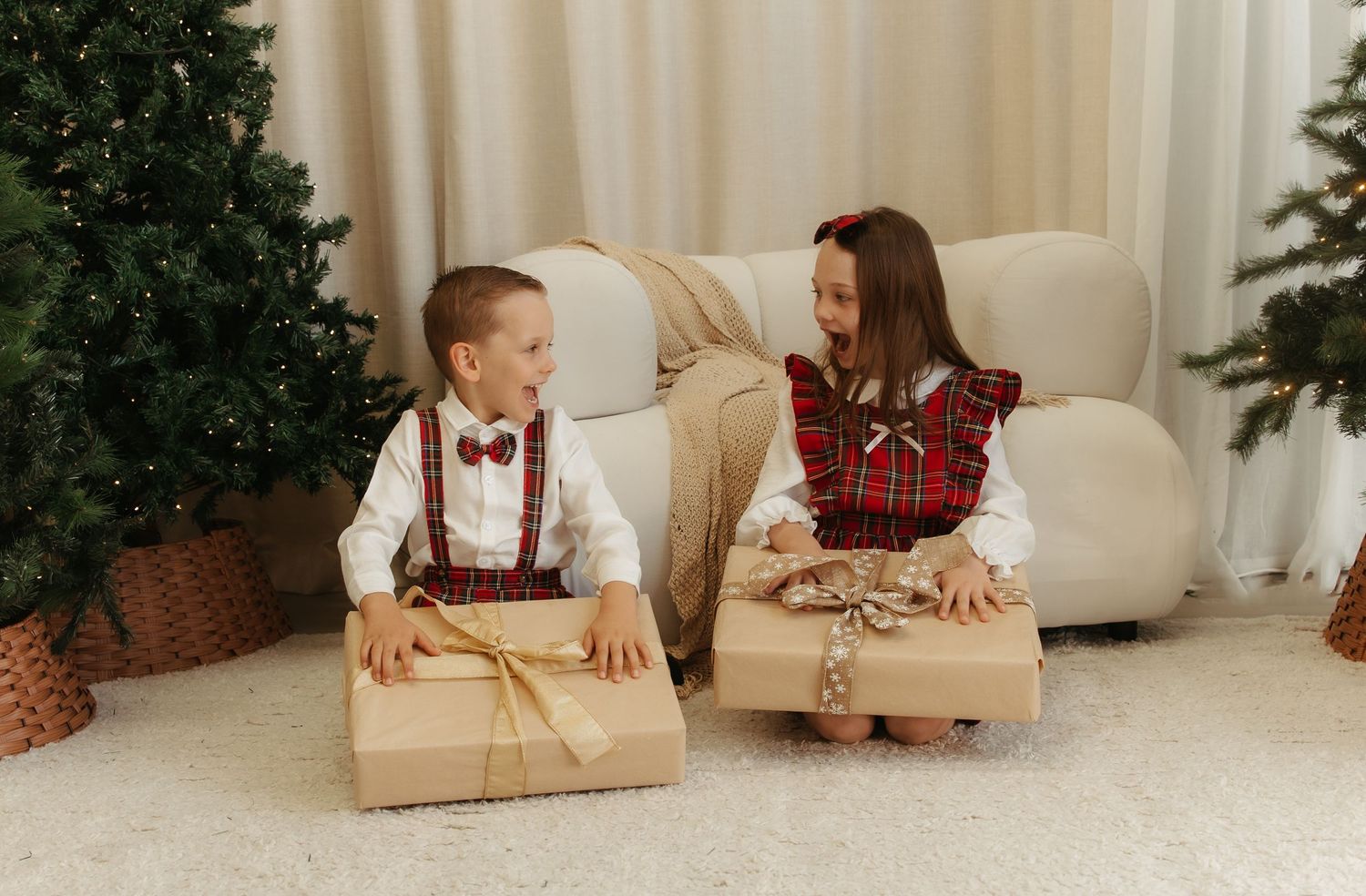 Two children in festive plaid outfits exchanging Christmas gifts near a decorated tree in a cozy living room.