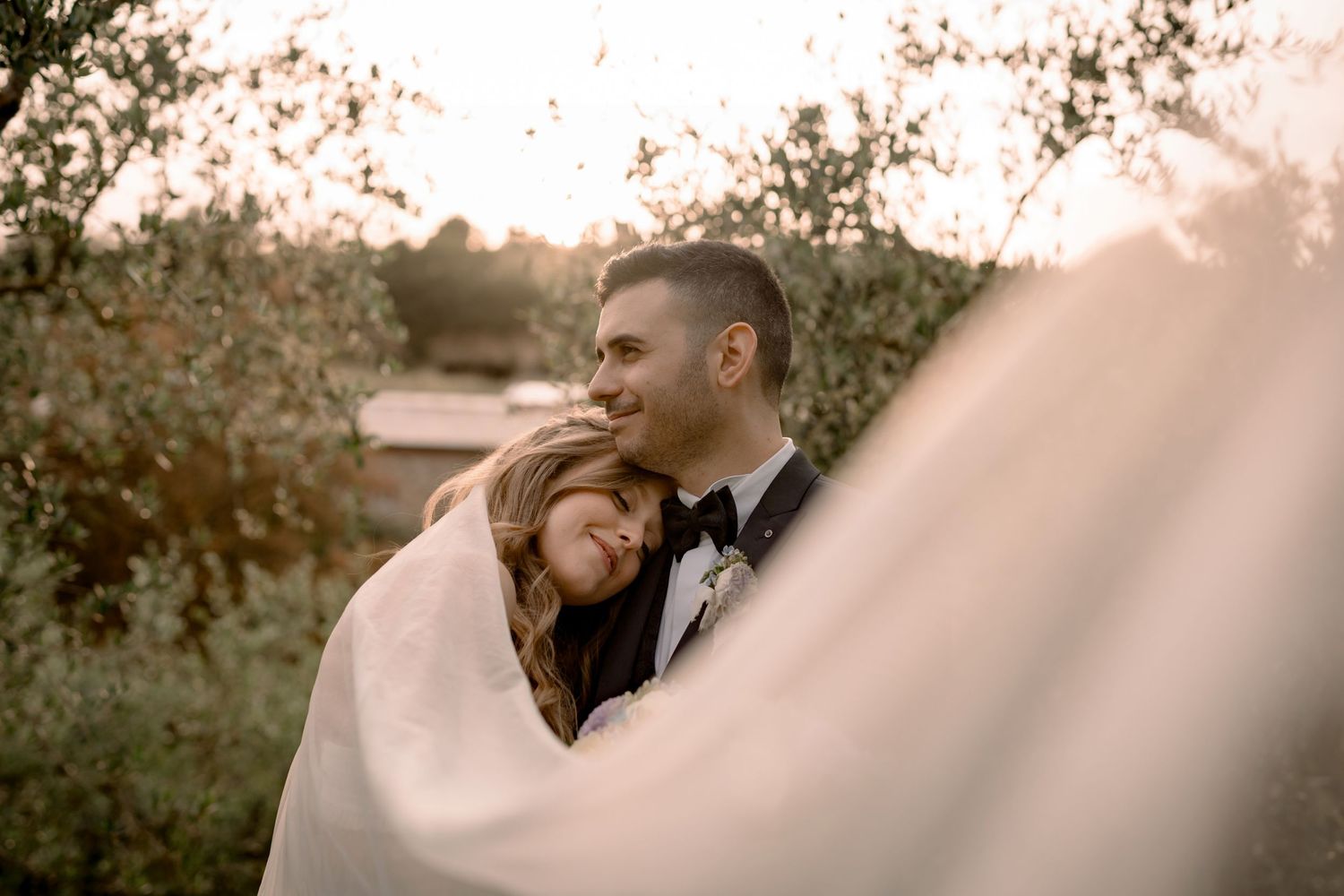 A bride and groom share a romantic embrace outdoors at golden hour, surrounded by lush greenery with a flowing veil.