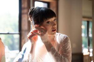 A bride in lace dress adjusts her earring, with soft natural light highlighting her elegant updo hairstyle.