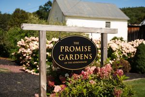 Sign for The Promise Gardens of the Adirondacks surrounded by pink flowering shrubs with a white barn in background.