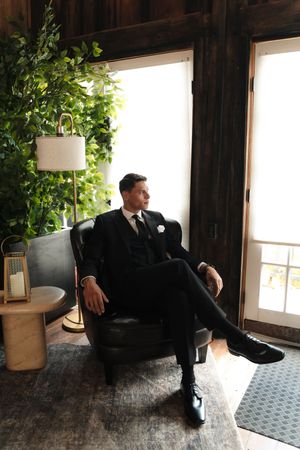 Groom in black suit sitting in leather chair by window with green plants and lamp in rustic wood room.