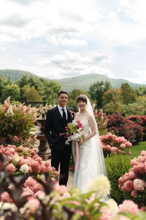 Bride and groom smiling among pink hydrangeas with mountain backdrop on their wedding day.