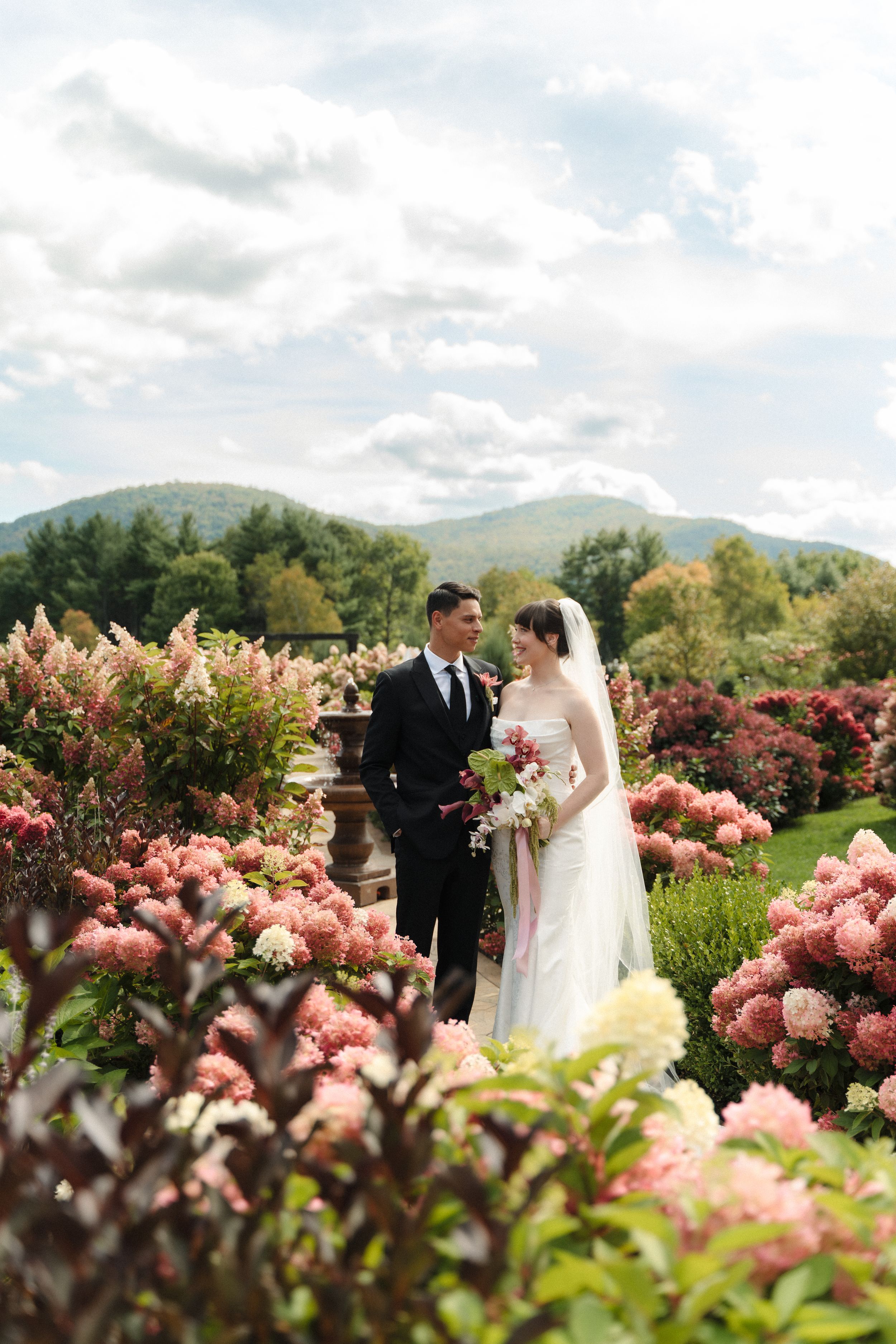 Bride and groom standing among pink hydrangeas with mountain views on their wedding day.