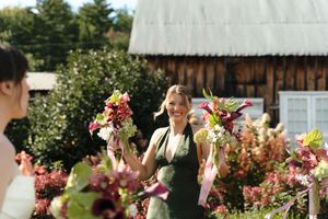 Smiling woman in green apron holding colorful flower bouquets outdoors near a rustic barn.