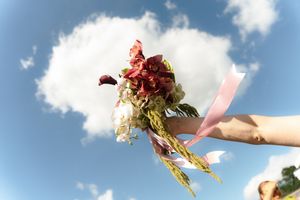 Hand holding a vibrant red and pink floral bouquet with ribbon against a blue sky with white clouds.