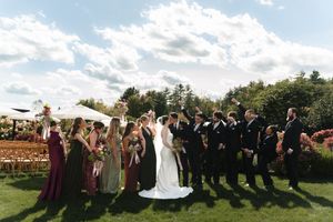 Wedding party poses outdoors on green lawn, bridesmaids in jewel-tone gowns and groomsmen in black suits under cloudy sky.