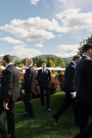 Groom stands confidently among groomsmen in dark suits on a green lawn with mountain views and blue sky.