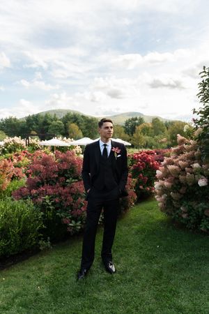 Groom in black suit with boutonniere stands in lush garden with colorful flowers and mountain backdrop.