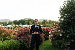 A young man in a navy suit stands in a lush garden with colorful blooming flowers and green hills in the background.