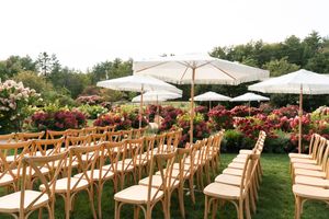 Outdoor wedding ceremony setup with wooden cross-back chairs, white umbrellas, and colorful floral garden backdrop.