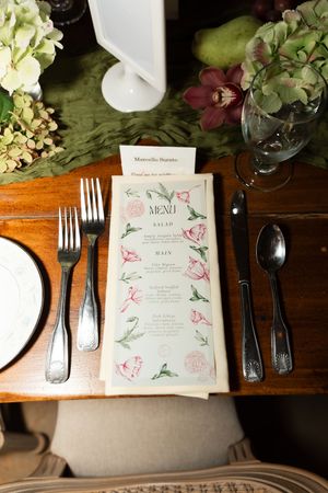 Wedding reception place setting with floral menu card, silver cutlery, and orchid floral centerpiece on wood table.