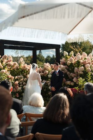 Outdoor wedding ceremony with bride and groom at floral altar surrounded by blush hydrangeas, guests seated under white tent.