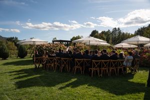 Guests seated at long outdoor dining tables on green lawn with white umbrellas and lush trees under blue sky.