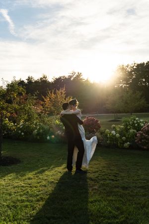 Groom lifting bride in white dress at golden sunset in lush garden during outdoor wedding celebration.