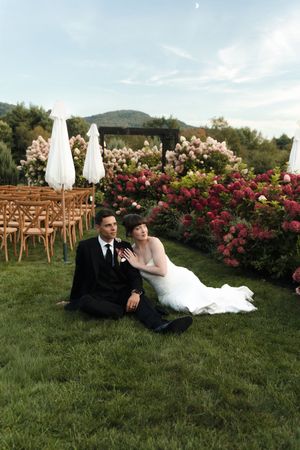 Bride and groom sitting on lush green grass beside floral ceremony arch and wooden chairs outdoors.
