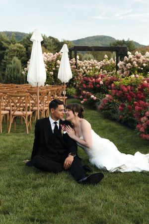 Bride and groom sitting together on lush green grass surrounded by pink hydrangeas at outdoor wedding venue.
