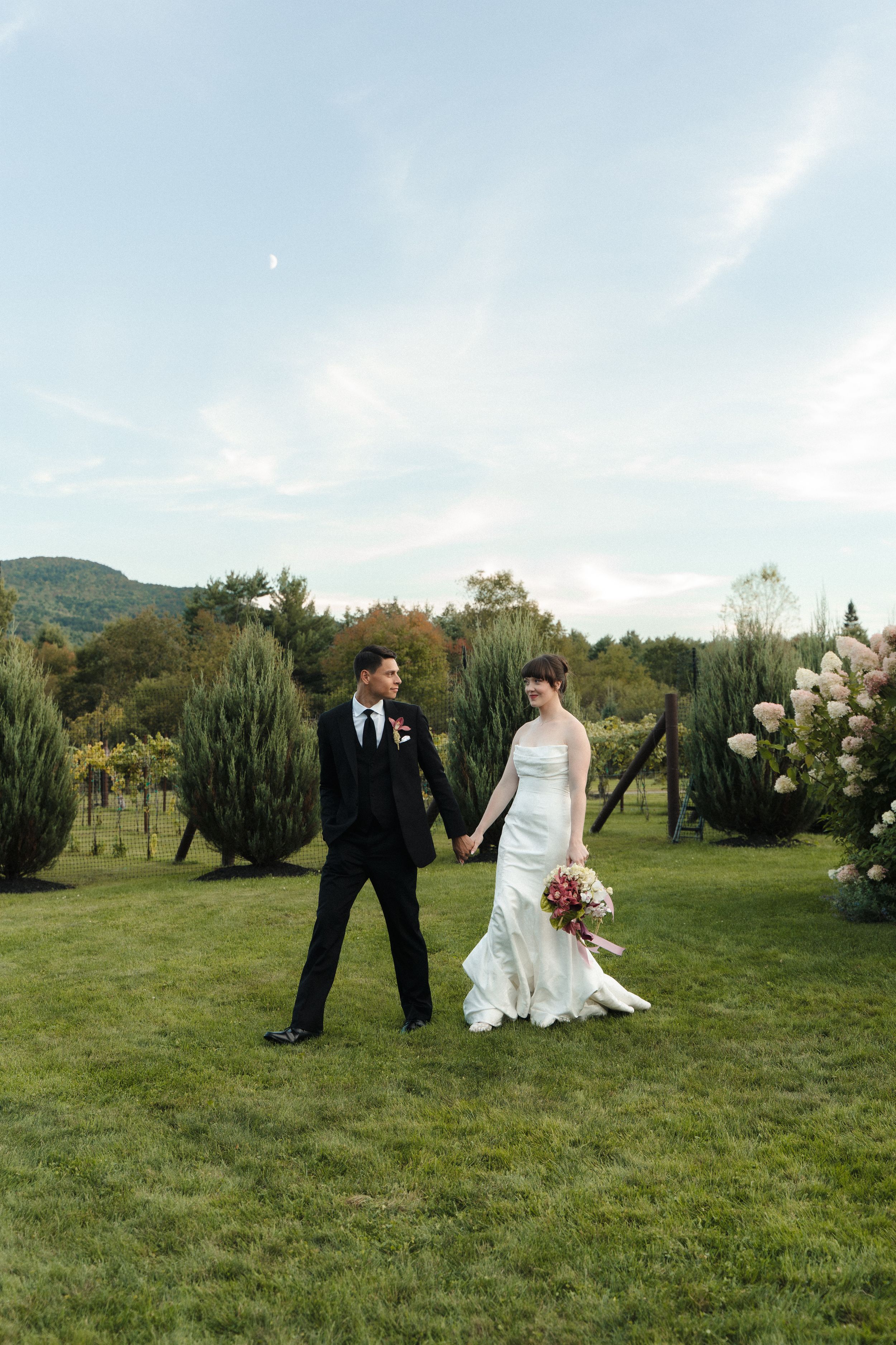 Bride and groom walk hand in hand across green lawn with mountain views on their wedding day.