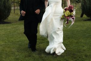 Bride in white gown holding colorful bouquet walks with groom in black suit on green grass at outdoor wedding.