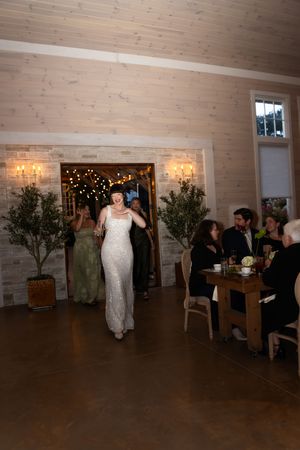 Bride in fitted white gown making a grand entrance into a warmly lit rustic barn reception hall.