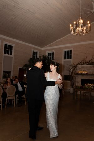 Bride in sequin gown and groom share first dance under chandelier in elegant rustic wedding venue.