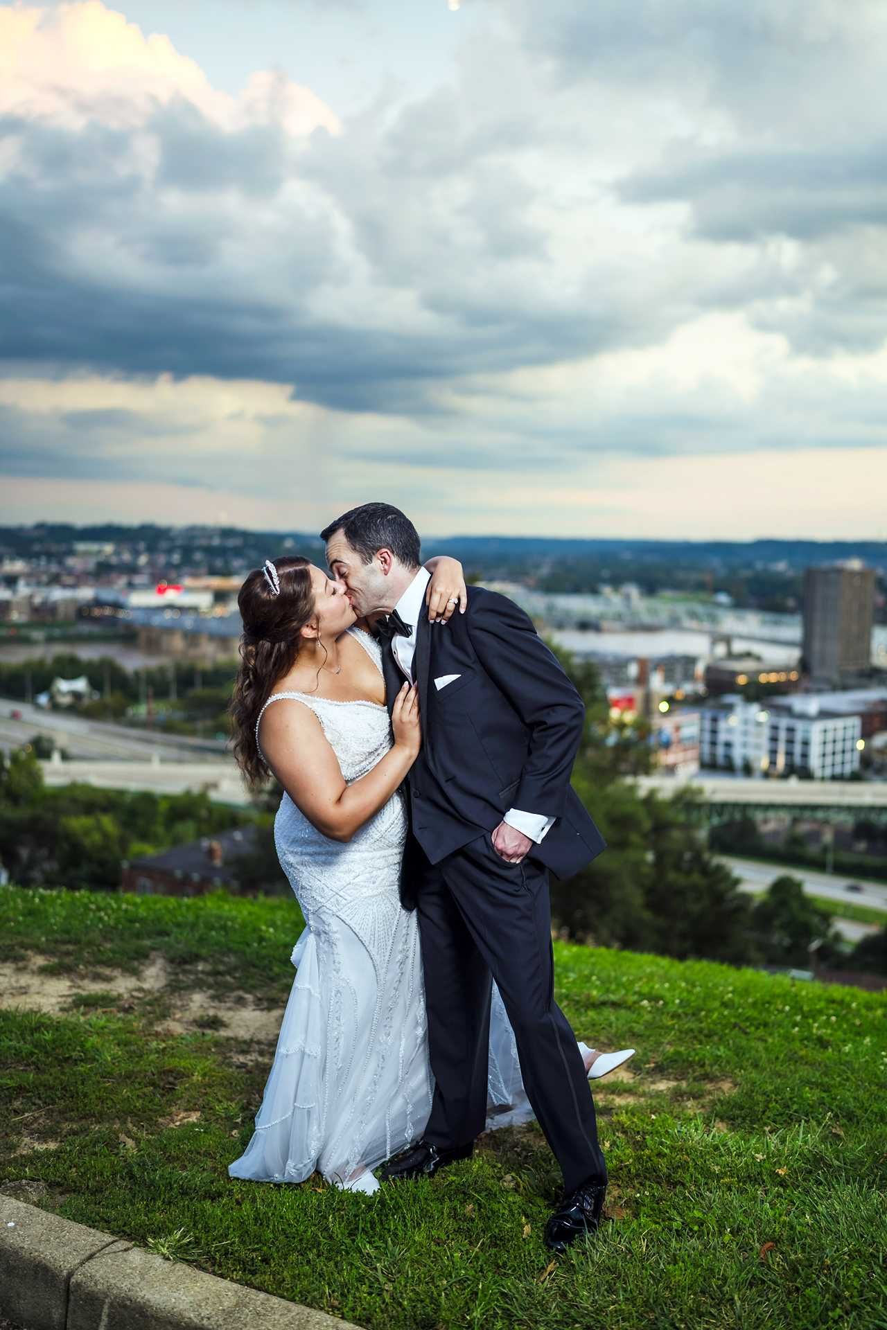 Bride and groom kissing on a grassy hill with a city skyline and dramatic cloudy sky in the background at the view wedding venue