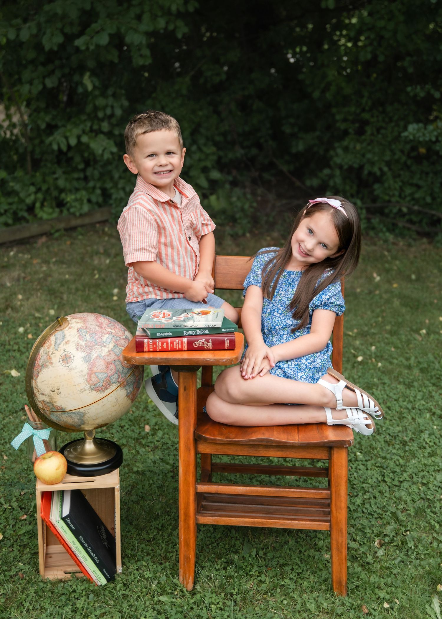 Two children pose outdoors with a vintage globe, books, and wooden school desk for a back-to-school portrait.