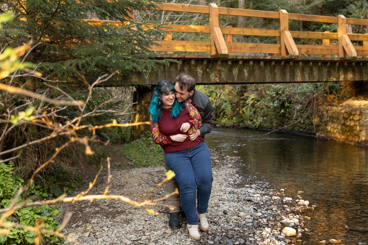 Couple embracing and laughing by a rocky creek with a wooden bridge surrounded by lush autumn foliage.
