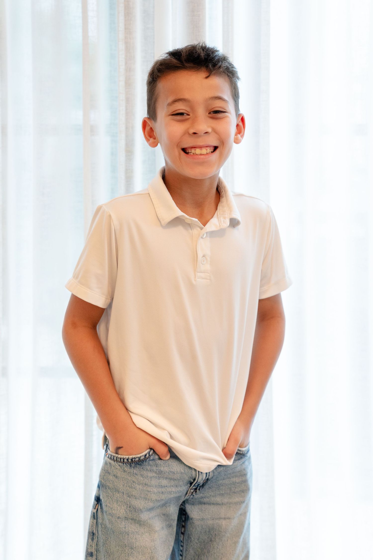 Smiling preteen boy in white polo shirt and jeans standing with hands in pockets against sheer curtains.