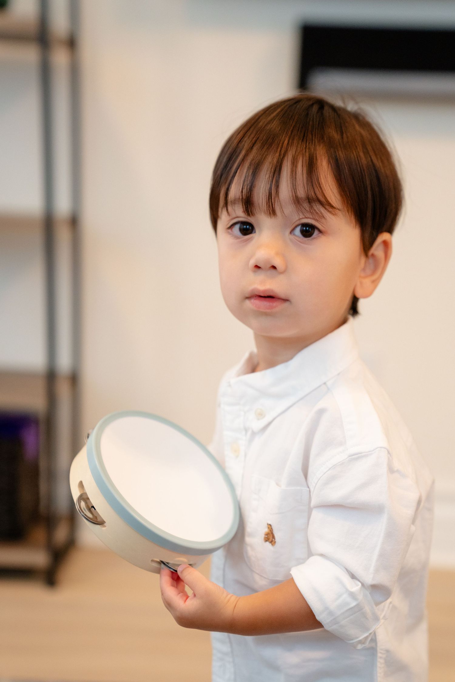 Young toddler boy in white shirt holding a small tambourine, looking at camera indoors.