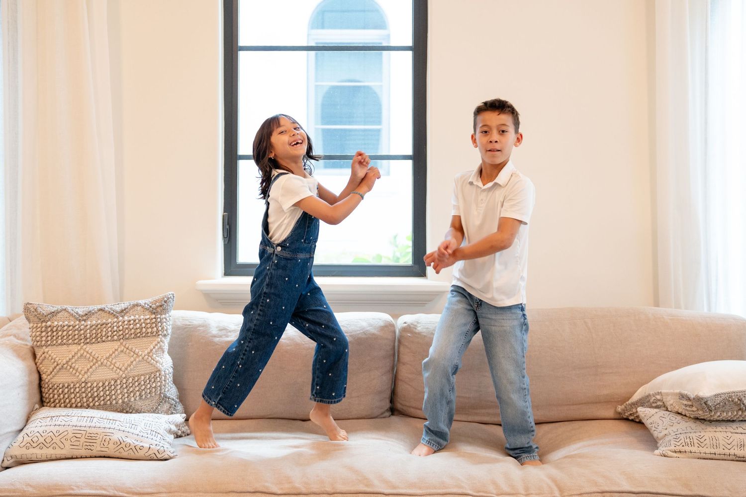 Two kids dancing and jumping on a couch in a bright living room, wearing casual denim outfits.