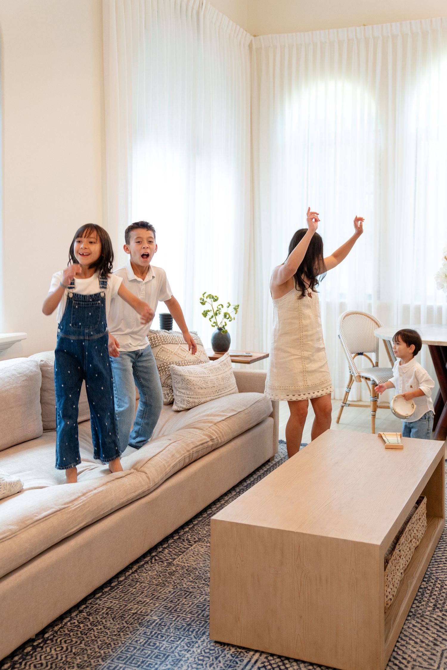 A joyful family with three kids and a mom jumping and dancing on a couch in a bright modern living room.