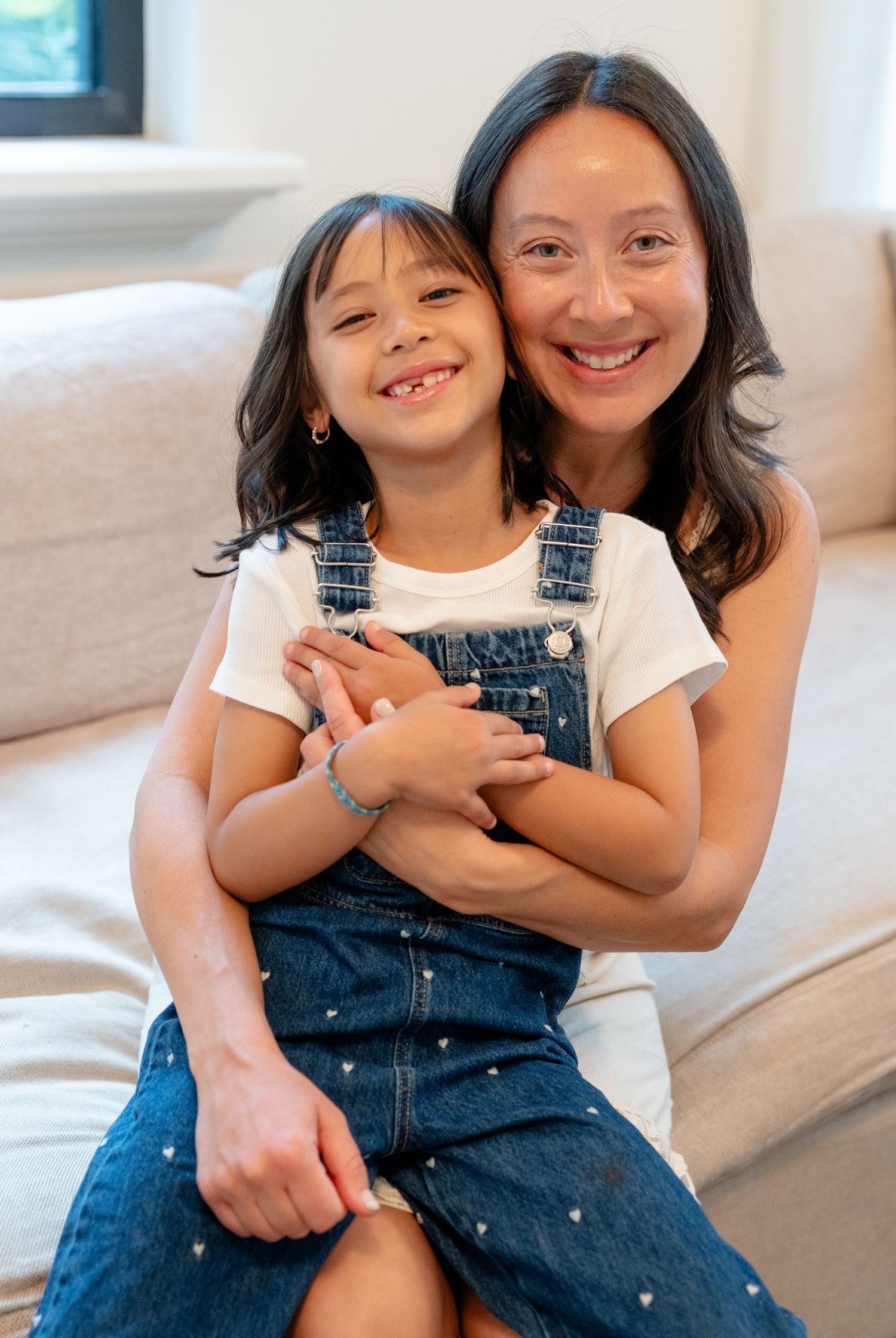 A smiling woman hugs a young girl in denim overalls on a couch.