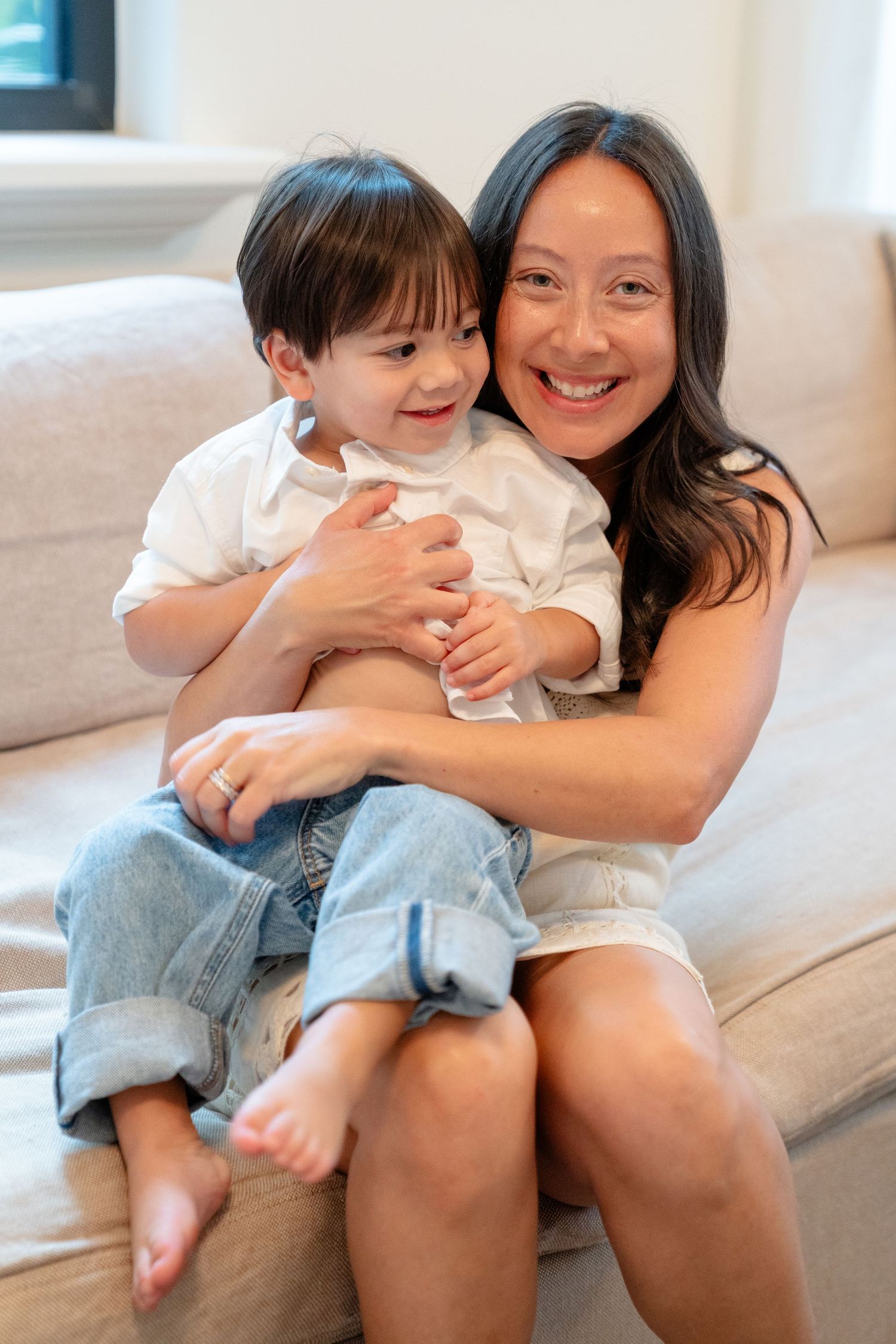 Smiling Asian mother hugging her young toddler son on a beige couch at home.
