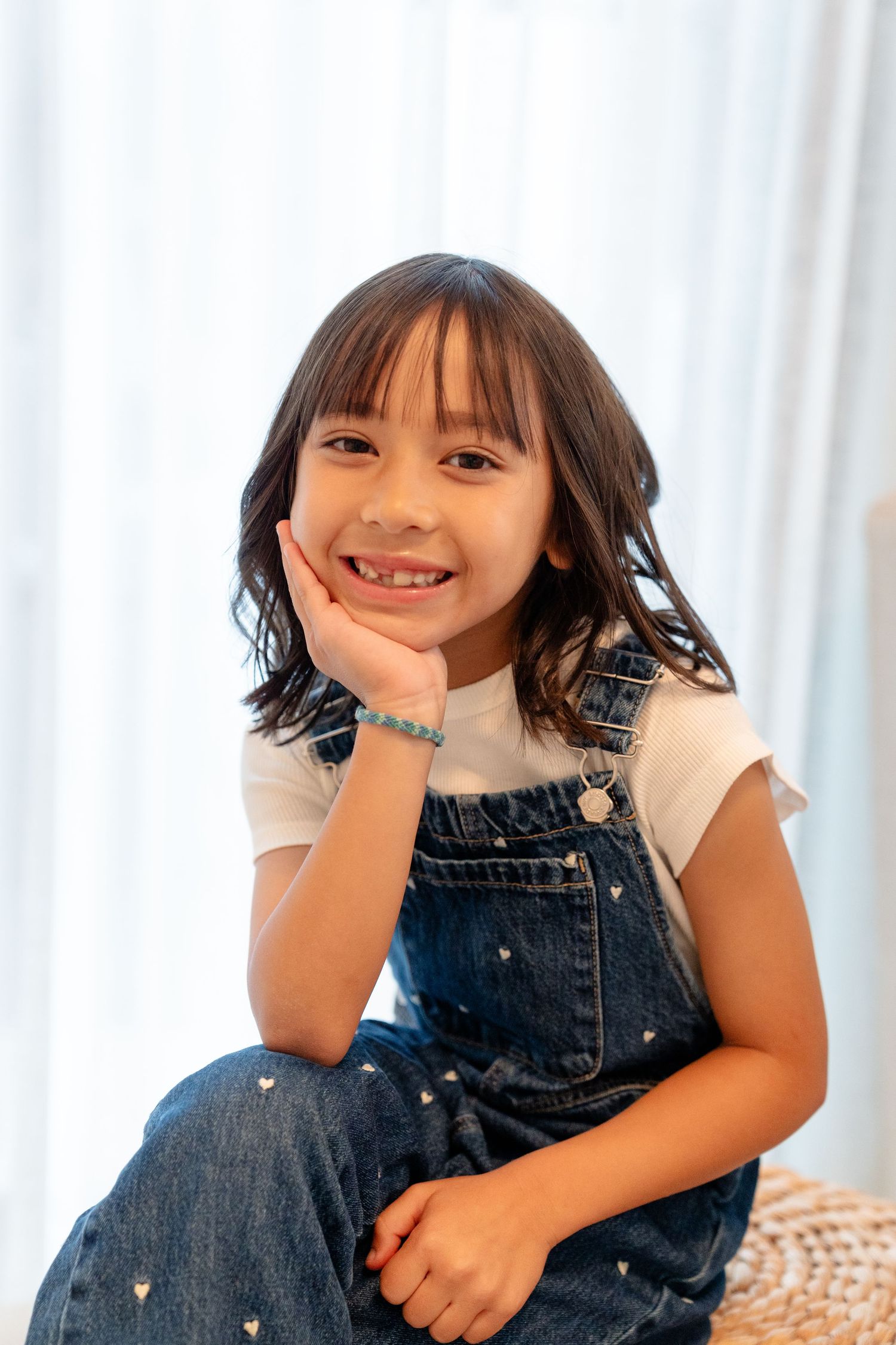 A smiling young girl with dark hair wearing denim overalls and a white shirt, resting her chin on her hand.