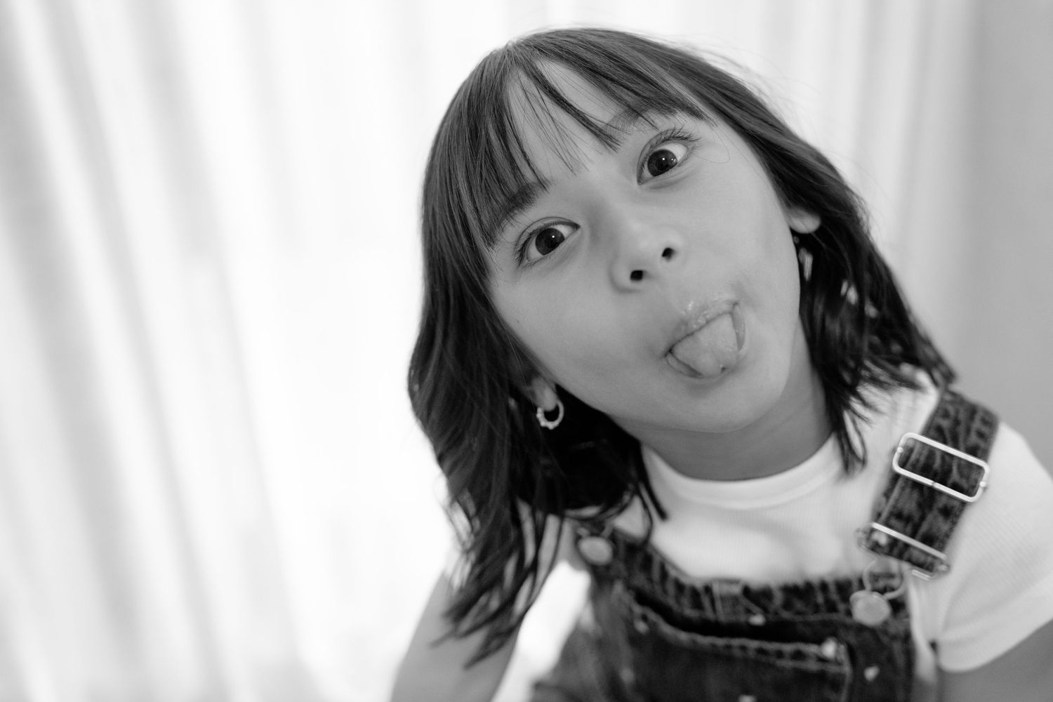Black and white photo of a young girl sticking her tongue out playfully, wearing overalls and a white shirt.
