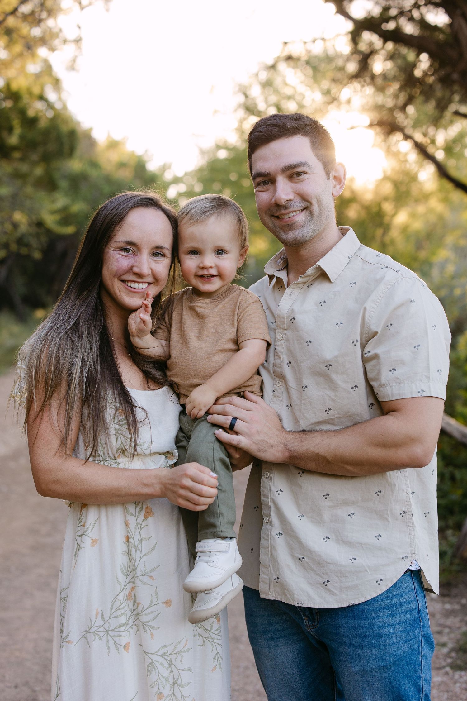 Family of five posing outdoors on a dirt path, dressed in white and neutral tones, smiling together under a tree.