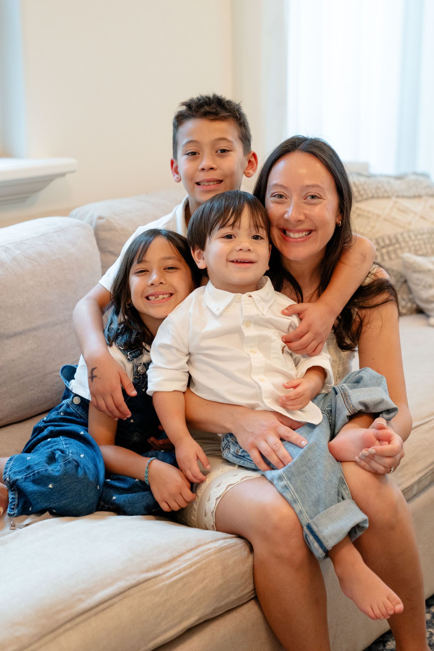 Smiling Asian mother sitting on couch with three happy children in casual home setting.