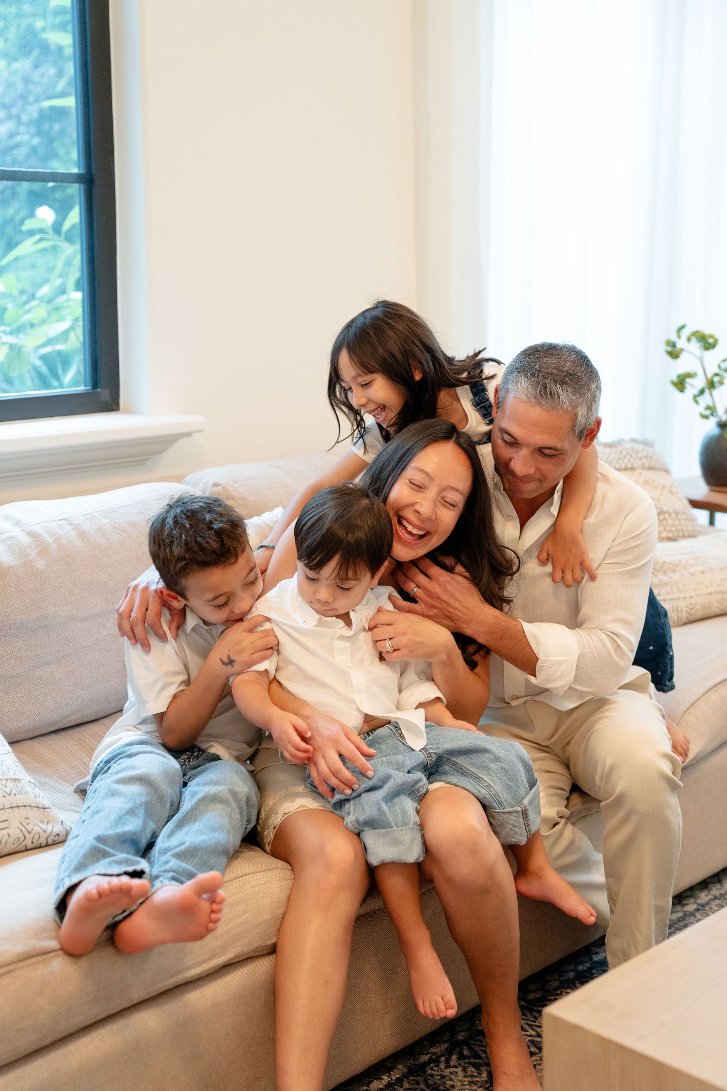 Happy Asian family of five laughing and cuddling together on a beige sofa in a bright modern living room.
