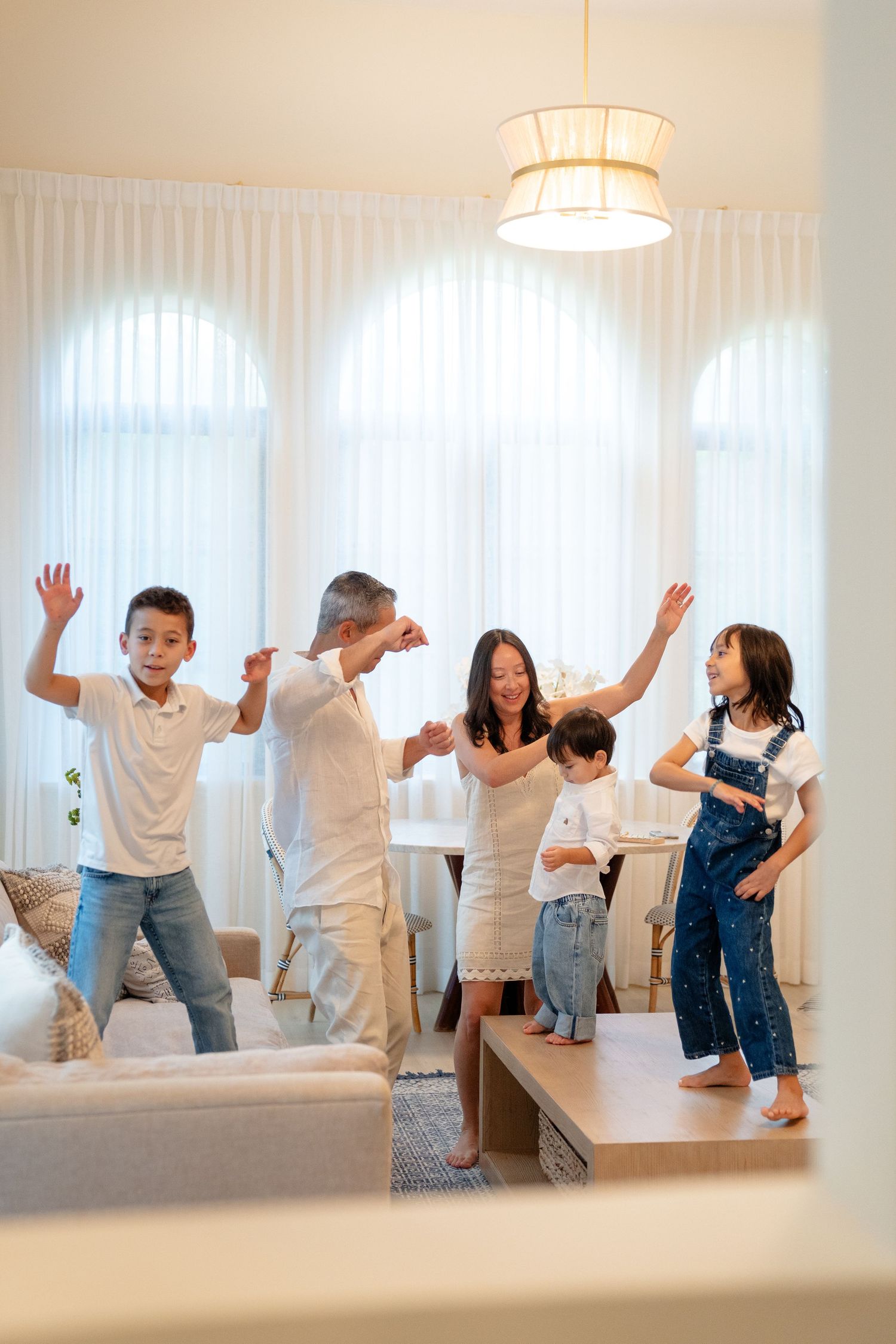 Asian family of five dancing and playing together in a bright modern living room with arched windows.